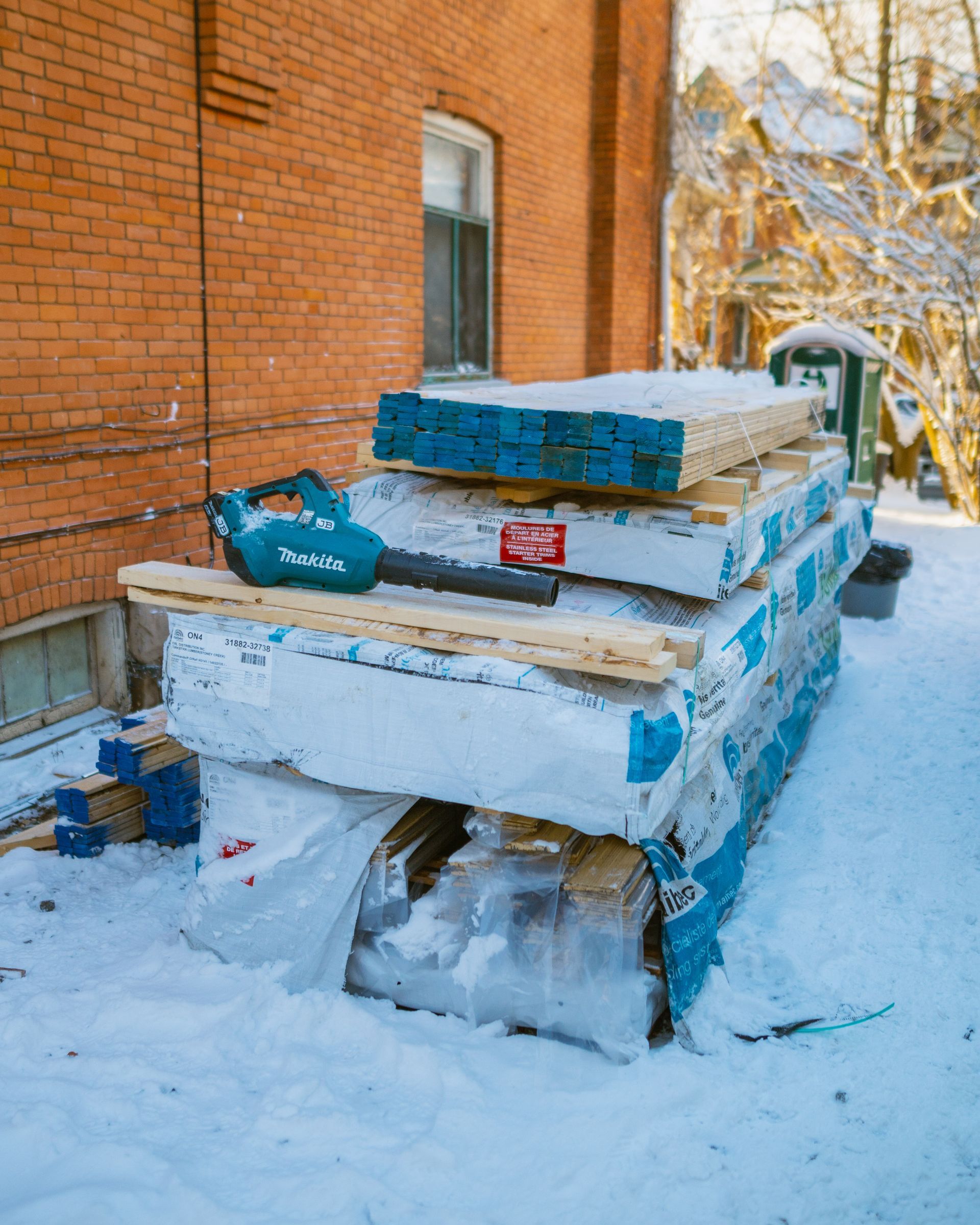 A pile of wood is sitting in the snow in front of a brick building.