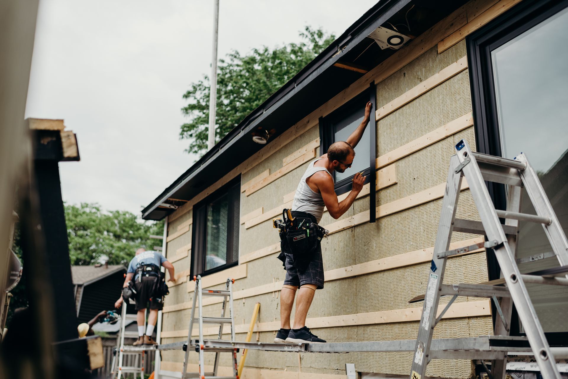 A man is standing on a ladder fixing a window on the side of a house.