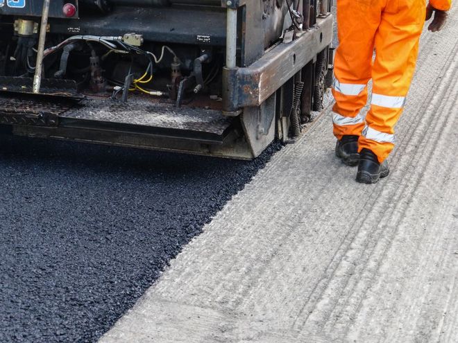 A road paver lays fresh black asphalt onto a textured gray road surface, with a worker in bright orange pants standing nearby.