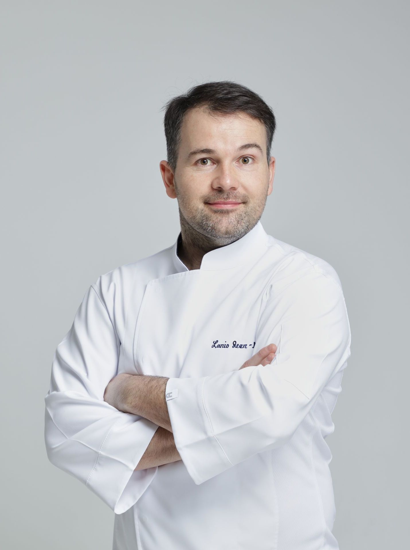 Un homme portant une veste de chef blanche est debout, les bras croisés.