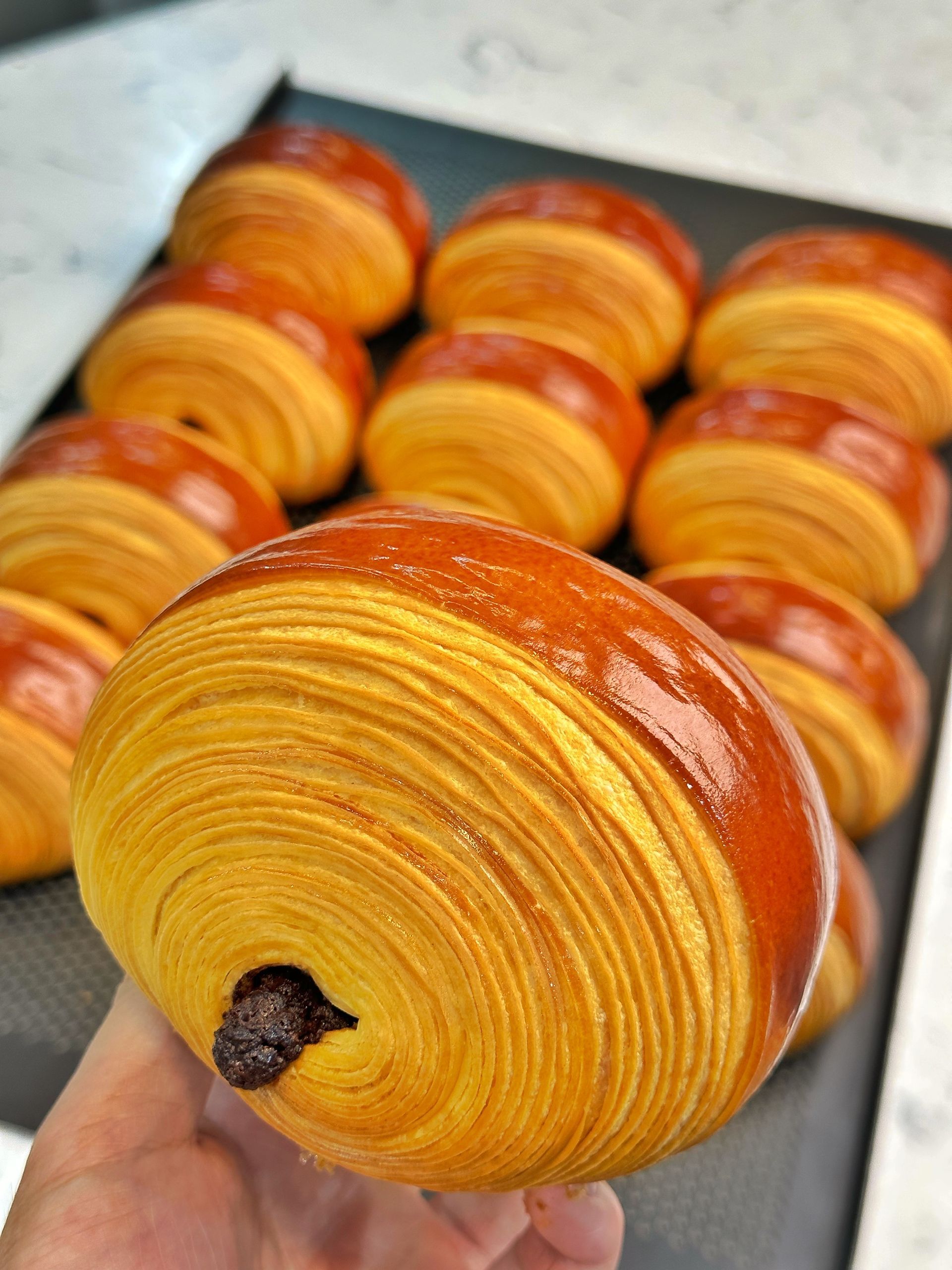 Une personne tient un croissant au chocolat devant un plateau de croissants