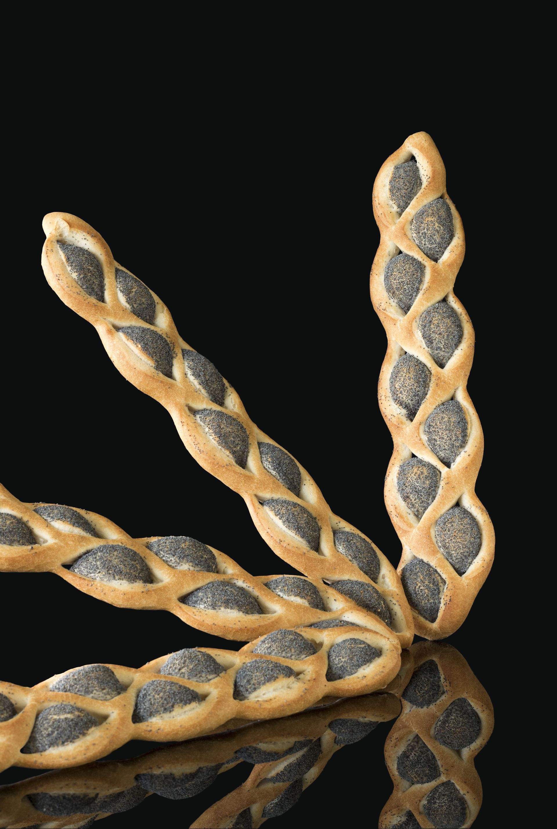 Trois bâtonnets de pain tressés avec des graines de pavot sur une surface noire.