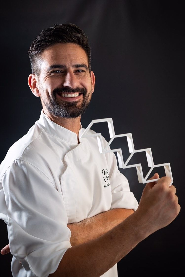 Un homme avec une barbe porte une veste de chef blanche et sourit.