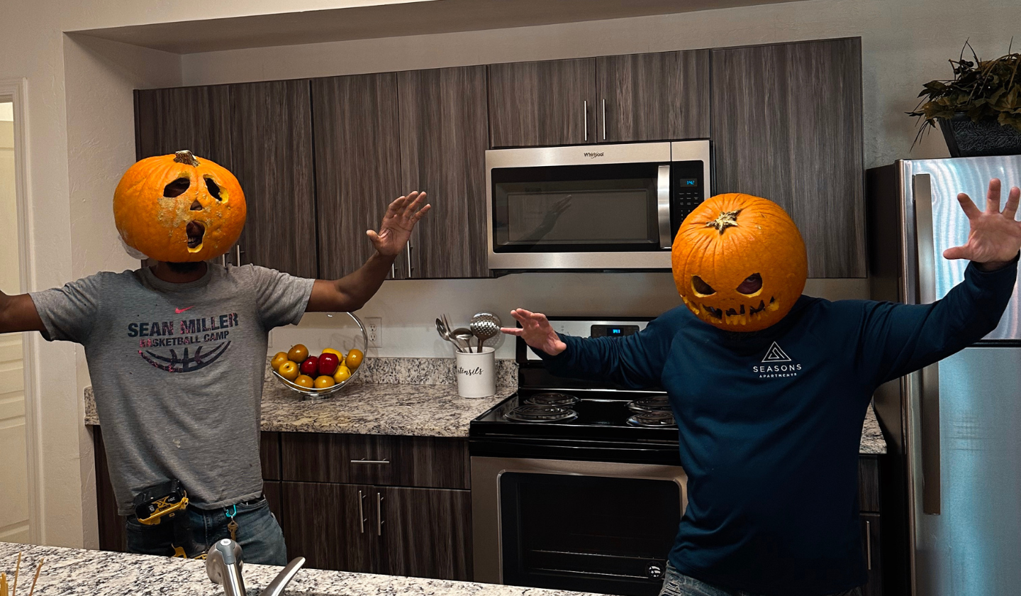 Two male residents in the kitchen wearing  carved pumpkin heads.
