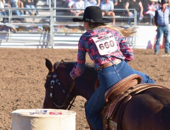 A female rider is on a brown horse during the Women's Barrel Racing event. Her long sleeved red and…