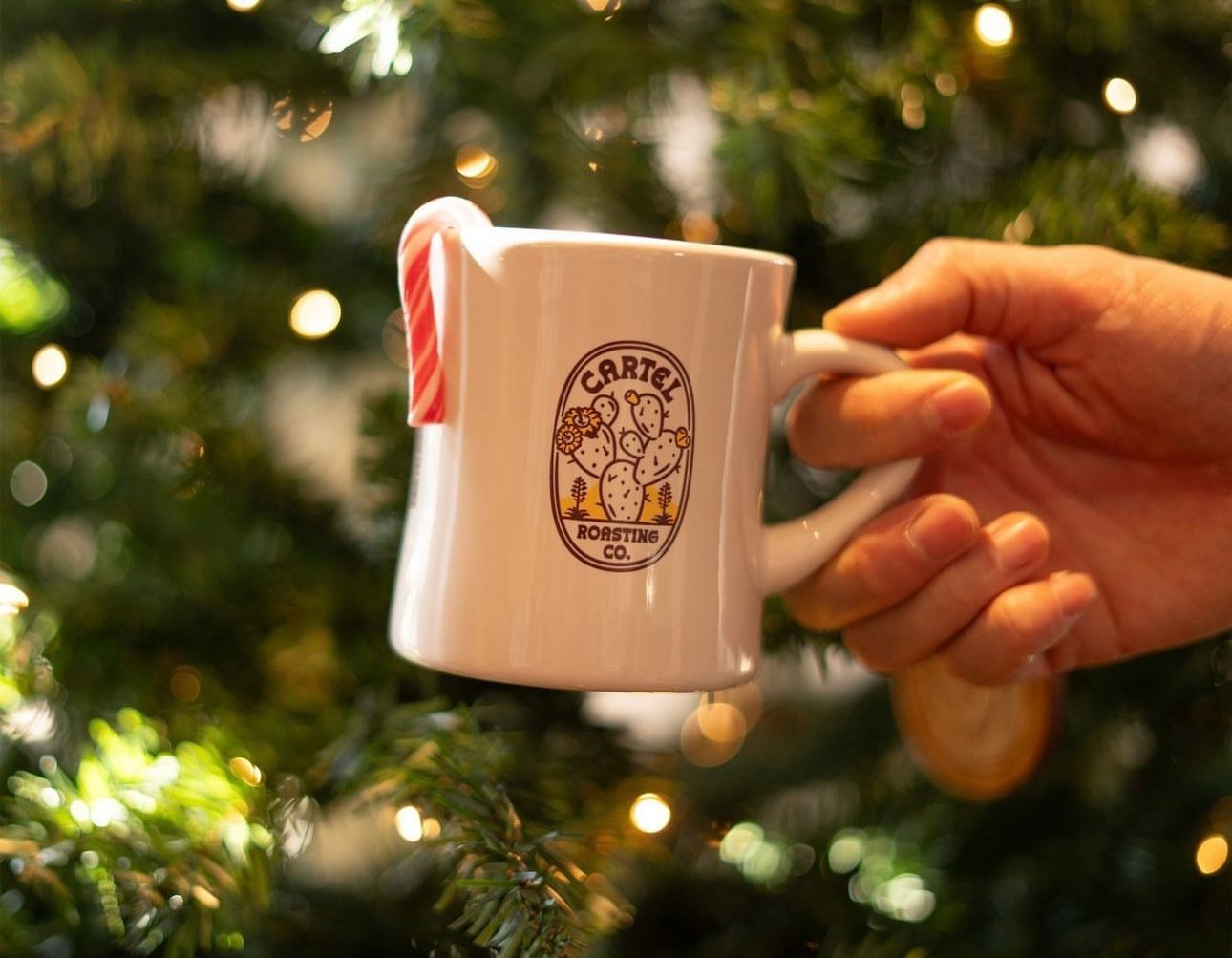  A festive mug with a candy cane, set against a twinkling Christmas tree from Cartel Roasting Co.