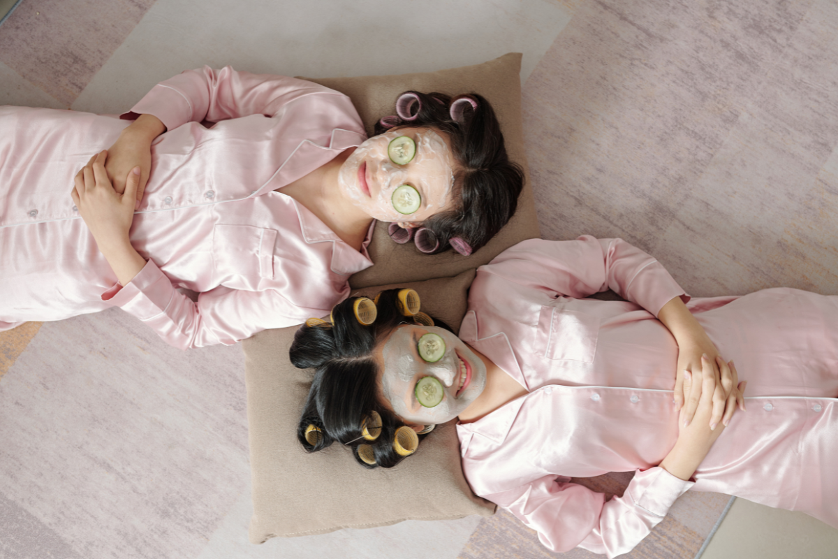 Two friends in pink pajamas, face masks, and curlers relax with cucumber eye masks.