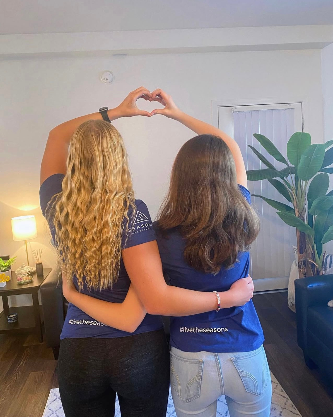 Two female students in blue logo shirts form a heart with their hands.