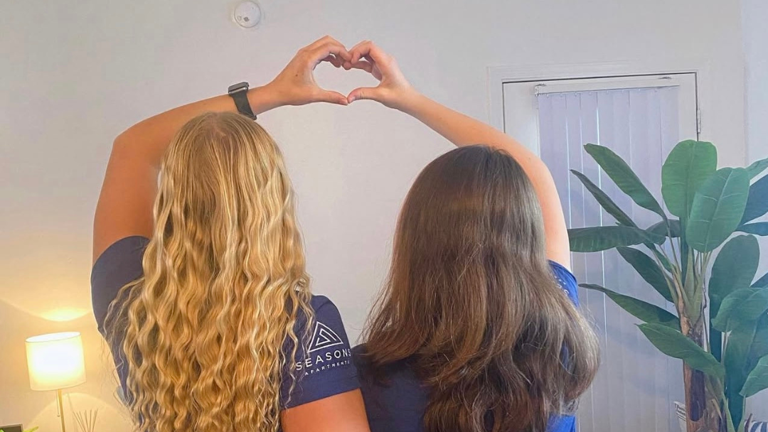 The backs of two female residents in their living room making a heart shape by joining their two hands above their heads.