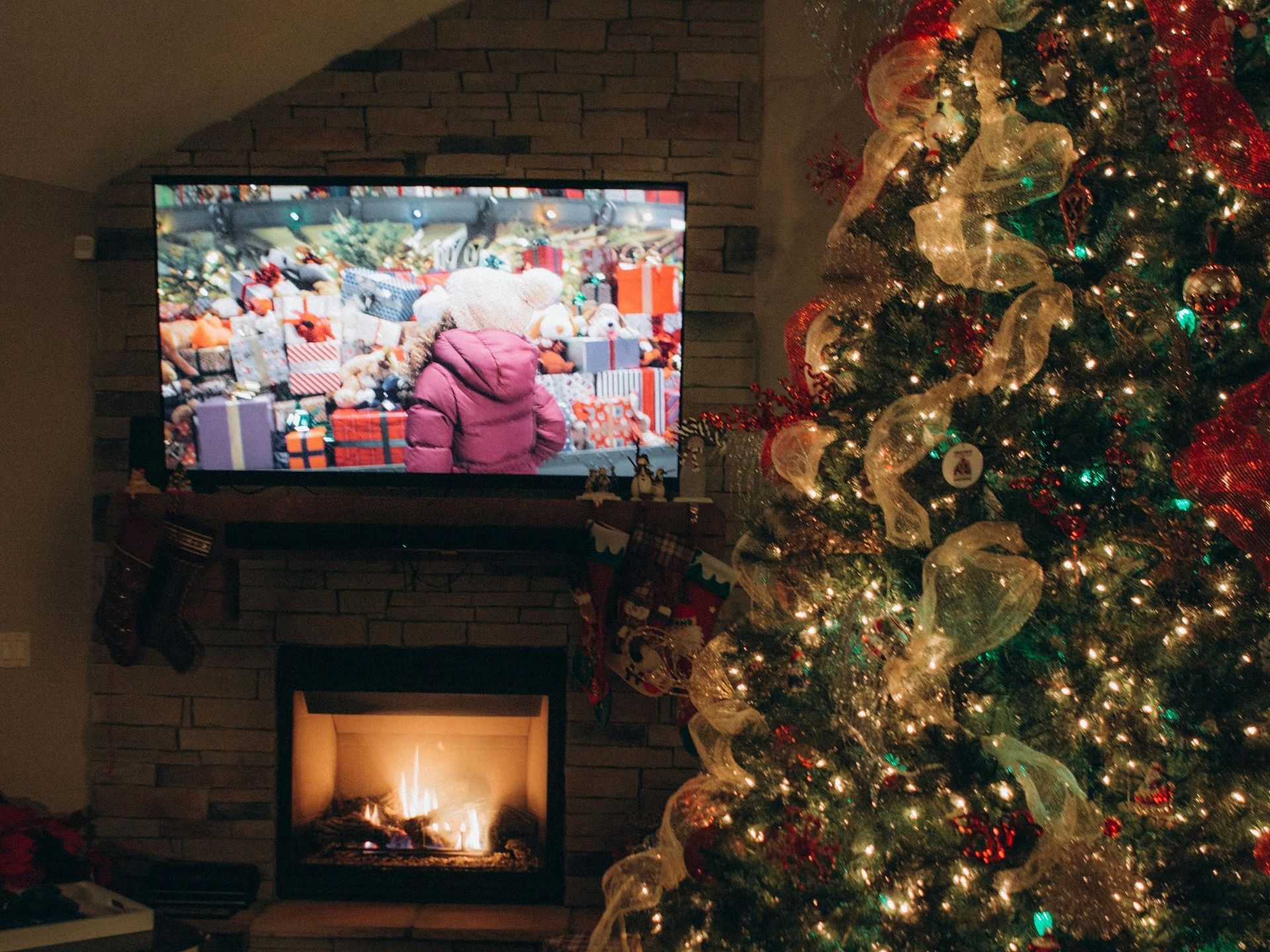 A living room decorated for Christmas with a Christmas tree, fire place, and a holiday movie playing on the big screen TV.