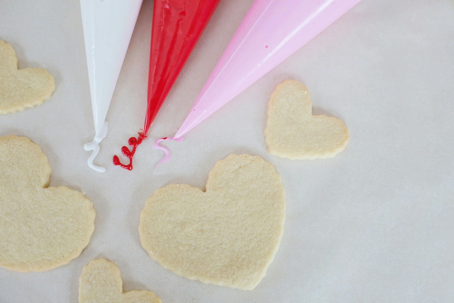 There are five heart shaped sugar cookies of various sizes. There are three icing tubes of the colors; red, white, and pink.