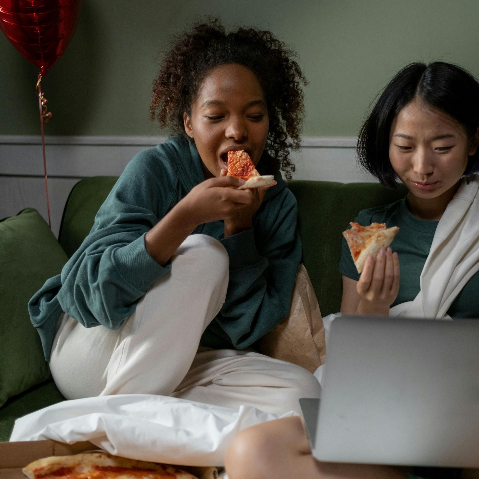 Two females are seen eating pizza and watching a laptop. They are sitting on a green couch with blankets.