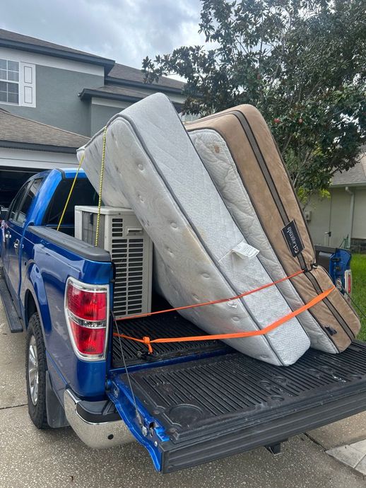 Blue pickup truck bed loaded with mattresses and an air conditioning unit, secured with orange straps.