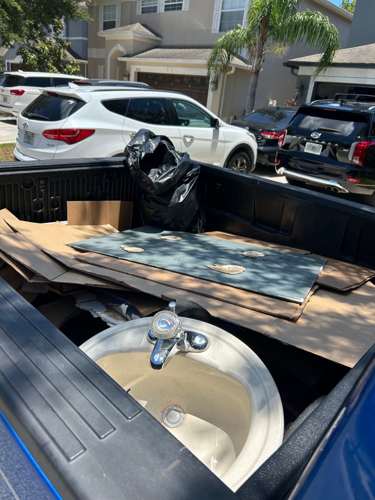 Pickup truck bed filled with renovation debris: sink, cardboard, boards, trash bag, parked cars, houses in background.