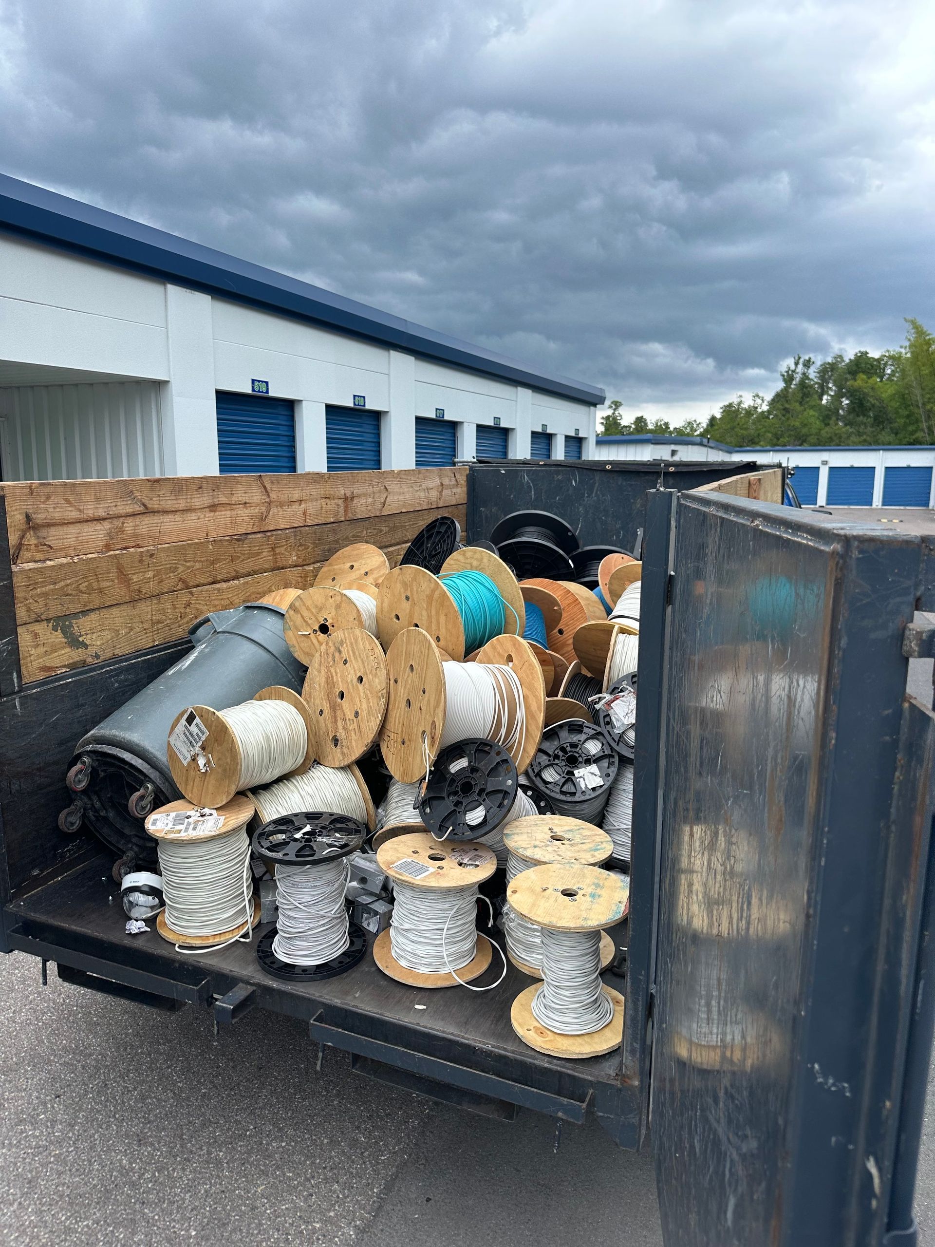 A truck bed filled with wooden spools of various colored wire and cables, parked in front of storage units.