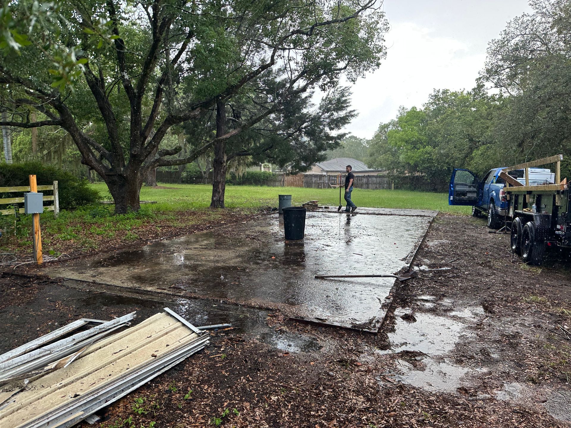 Muddy yard with a person, debris, and vehicles on a concrete slab in the rain.