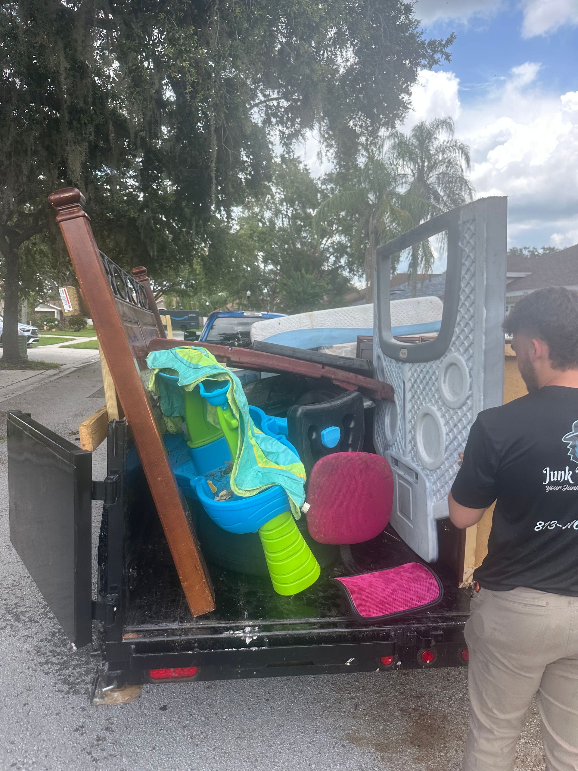 A person loads a trailer with debris. Items include a board, tub, towels, and a pink rug.