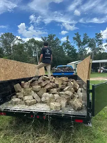 Person standing in a trailer loaded with bricks, with trees and cloudy sky in the background.