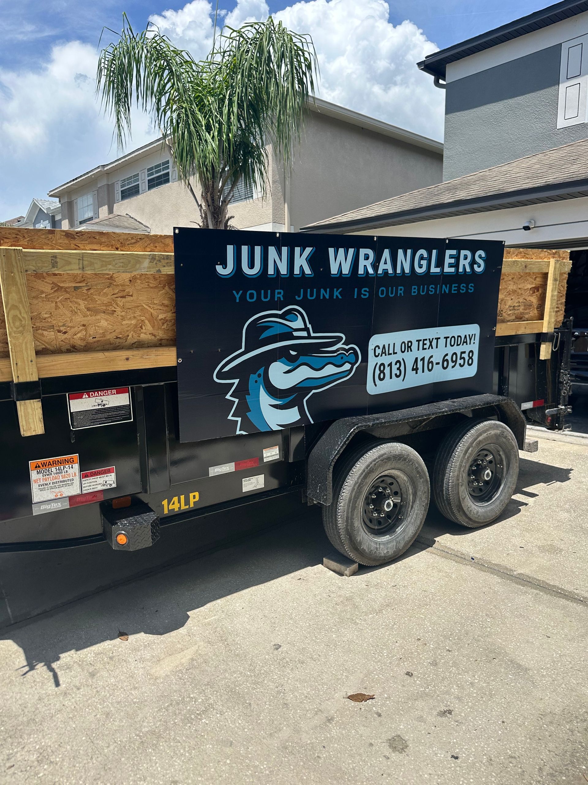 Junk Wranglers trailer with black logo, alligator mascot, and phone number in front of residential buildings.