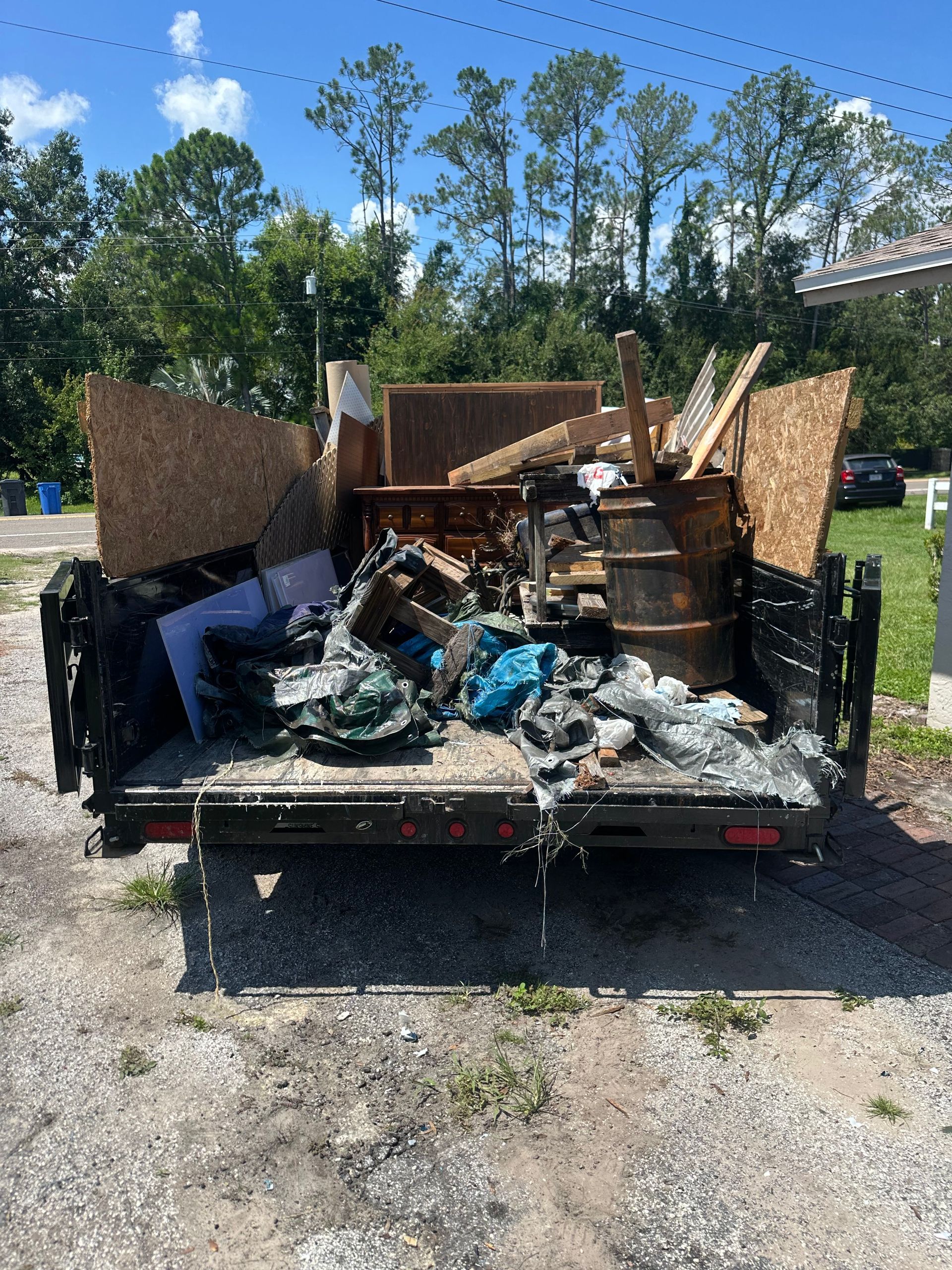 A black trailer filled with debris, including wood, a barrel, and other trash, parked outdoors.