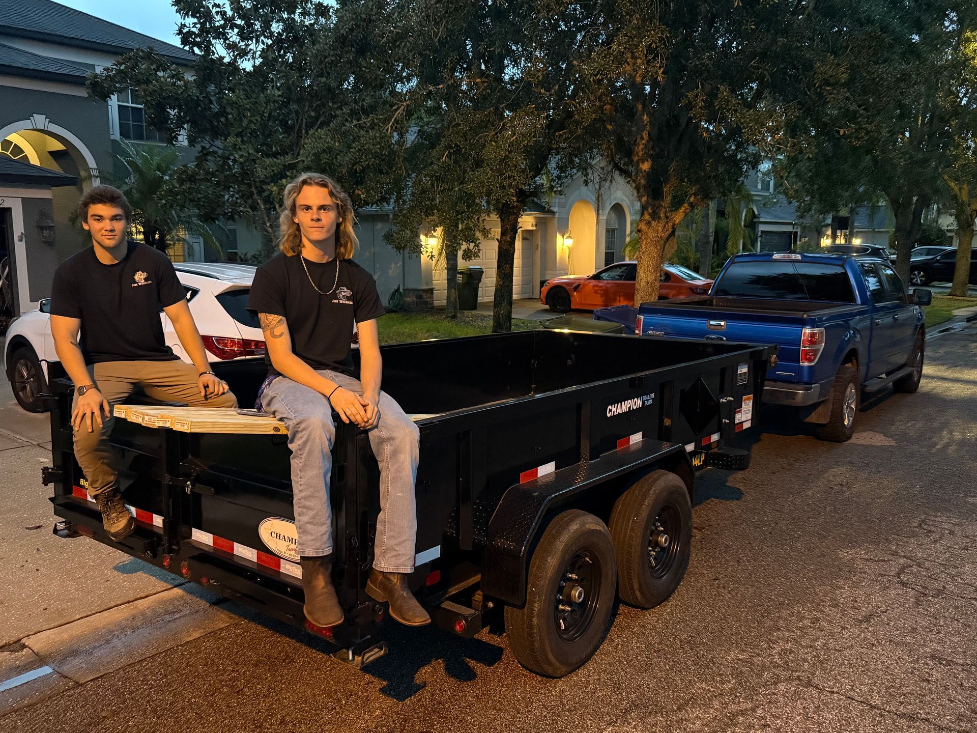 Two men sitting on a black trailer, parked on a street in front of a house.