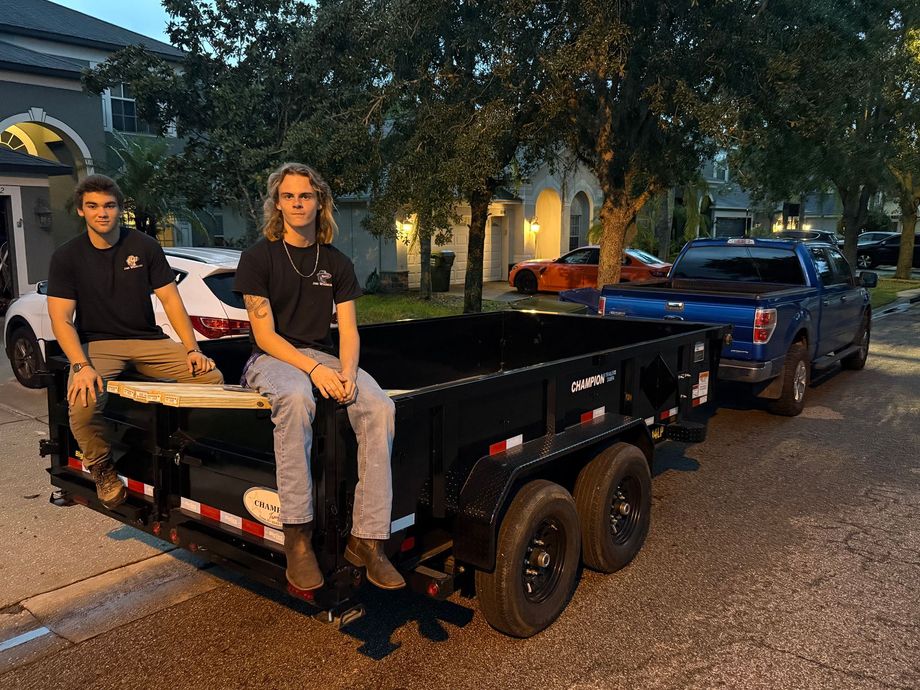 Two young men sit on a black trailer, parked on a street near houses. Blue truck and white car are nearby.