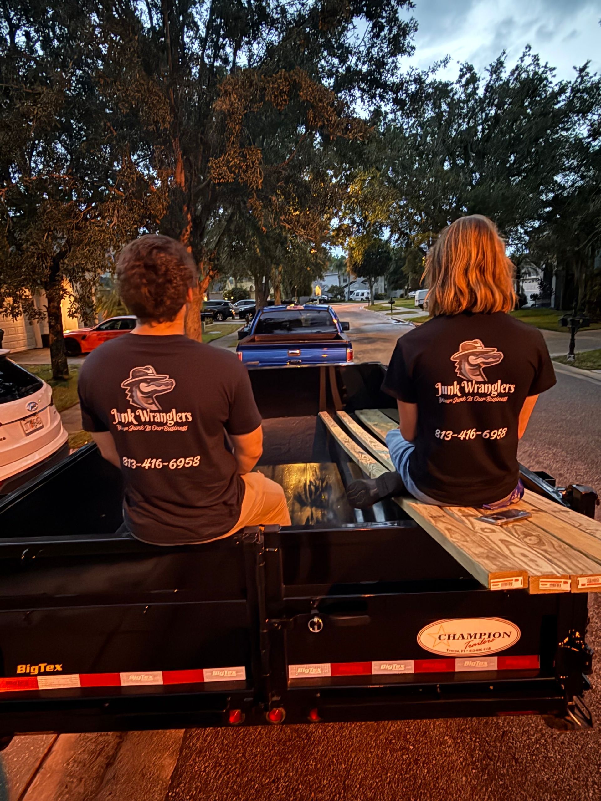 Two people in matching shirts sit in a trailer. The shirts read “Truck Wranglers” with a phone number.