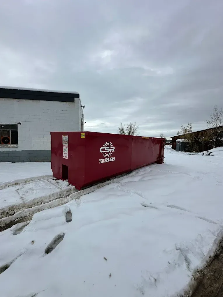 Red dumpster in snowy area next to a white building under a cloudy sky.