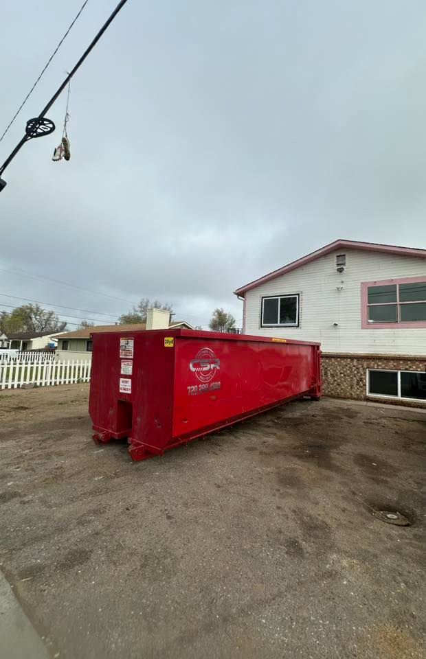Red dumpster in front of a white house on a cloudy day.