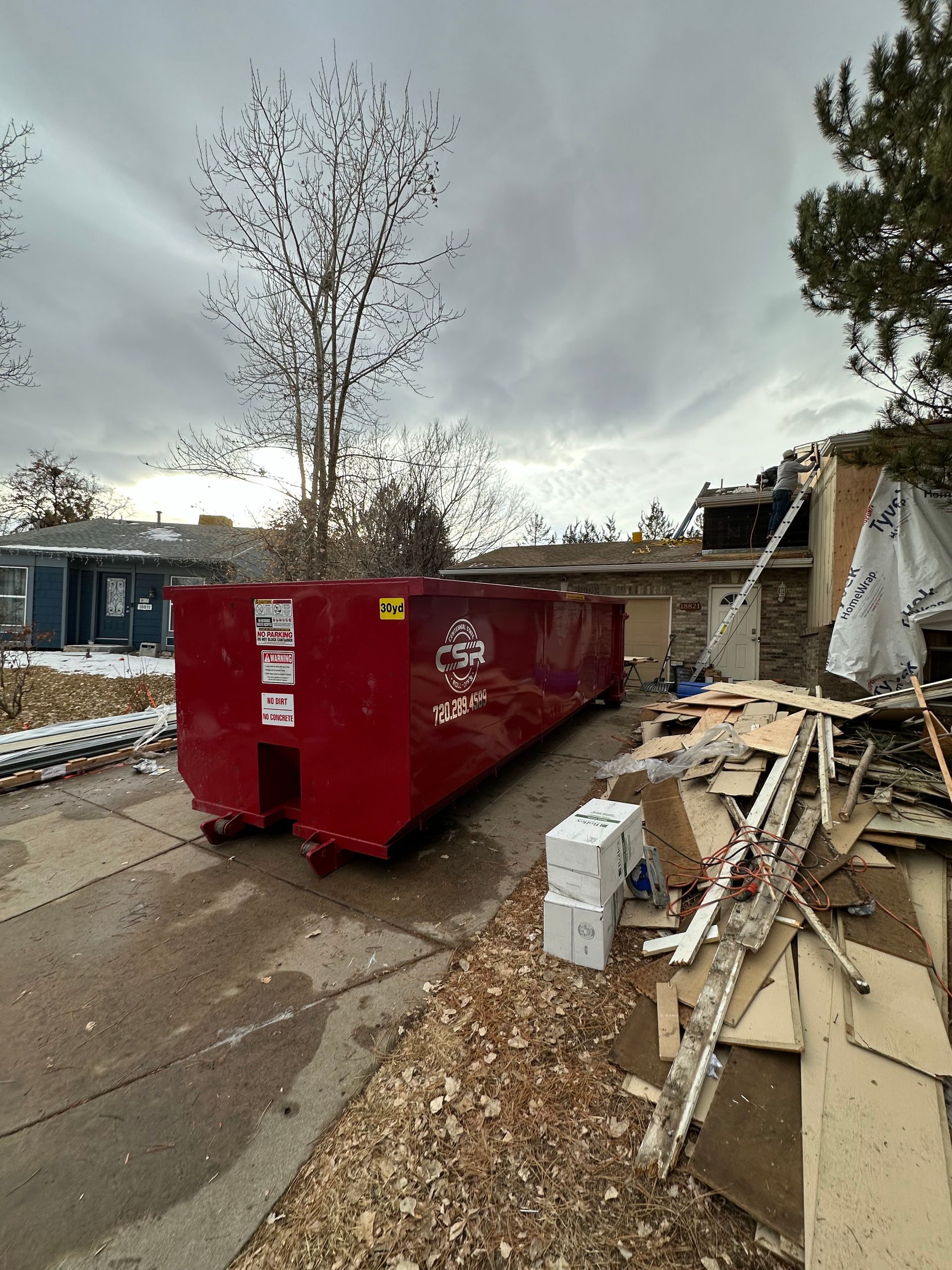 Red dumpster on a driveway next to construction debris and a house under repair. Cloudy sky.