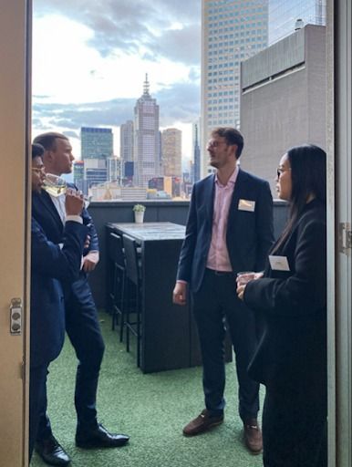 Three professionals in business attire socialize on a balcony overlooking a city skyline at dusk.