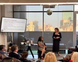A presenter stands next to a screen at a workshop, while another person sits in a chair during an indoor group session.
