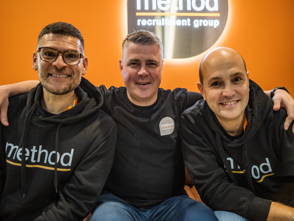 Three people smiling in black hoodies with the Method logo, posing in front of an orange Method Recruitment Group sign.