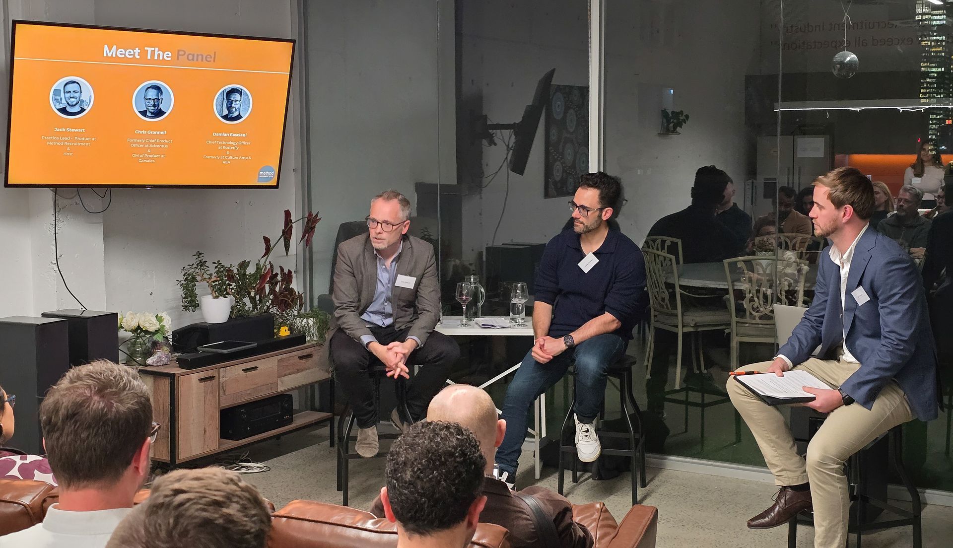 Three men in black hoodies smiling, posing in front of an orange wall with the 