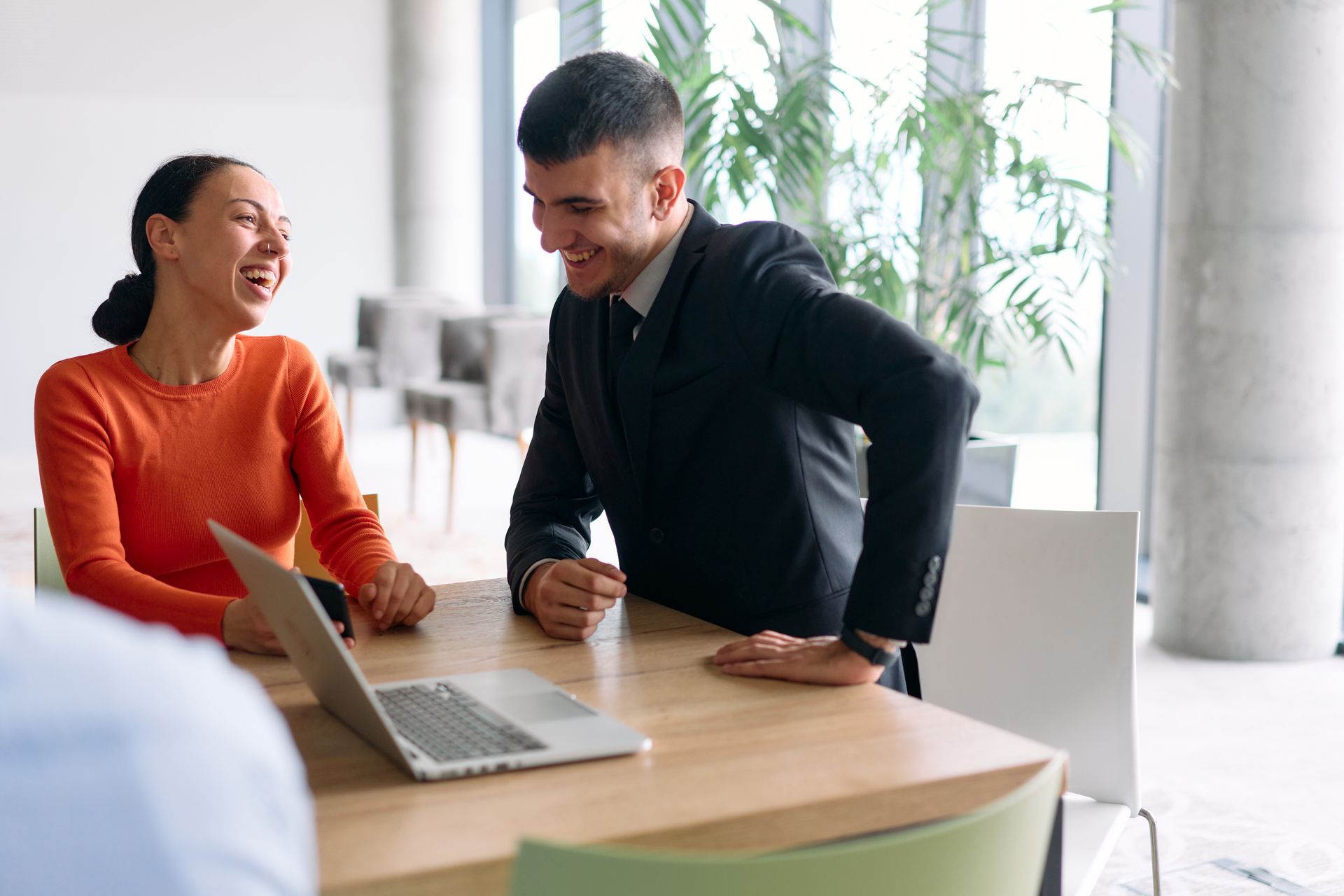 A person in a suit and a person in an orange sweater laugh while looking at a laptop together at a wooden office table.