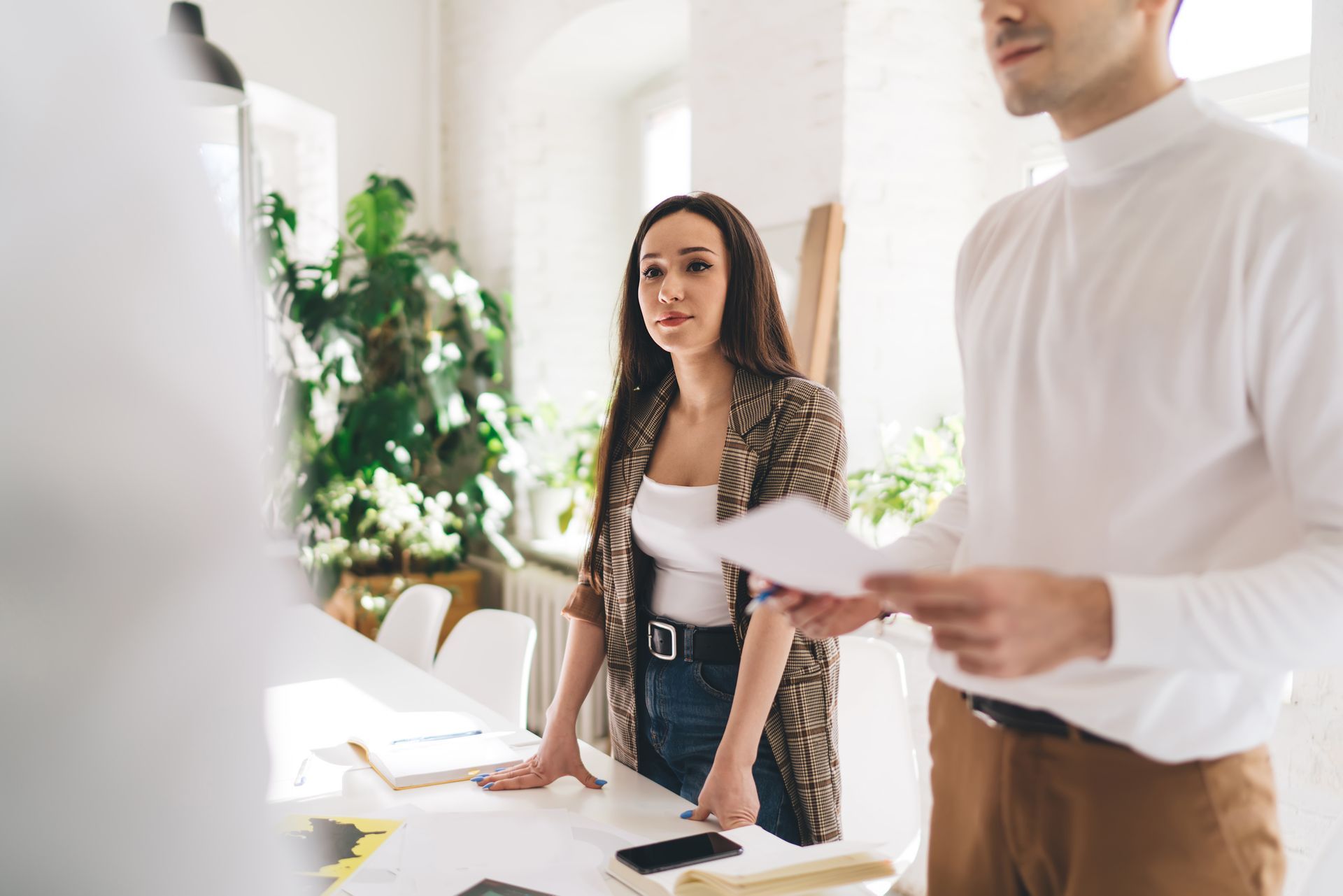Two people discussing designs at a table in a bright workshop; woman looking thoughtful, man holding papers.