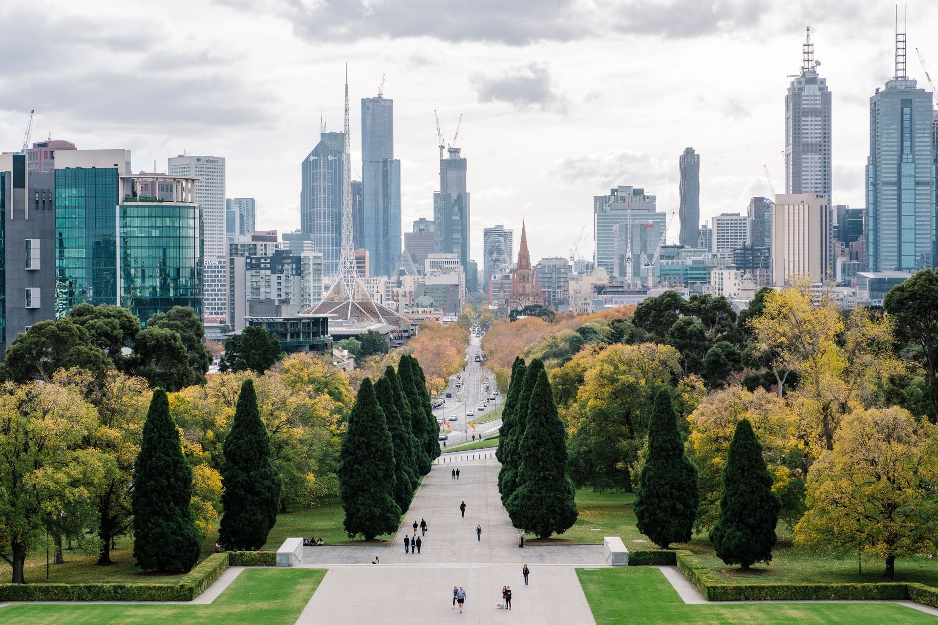 A city skyline with tall buildings is seen from a park with a tree-lined avenue and walking people.