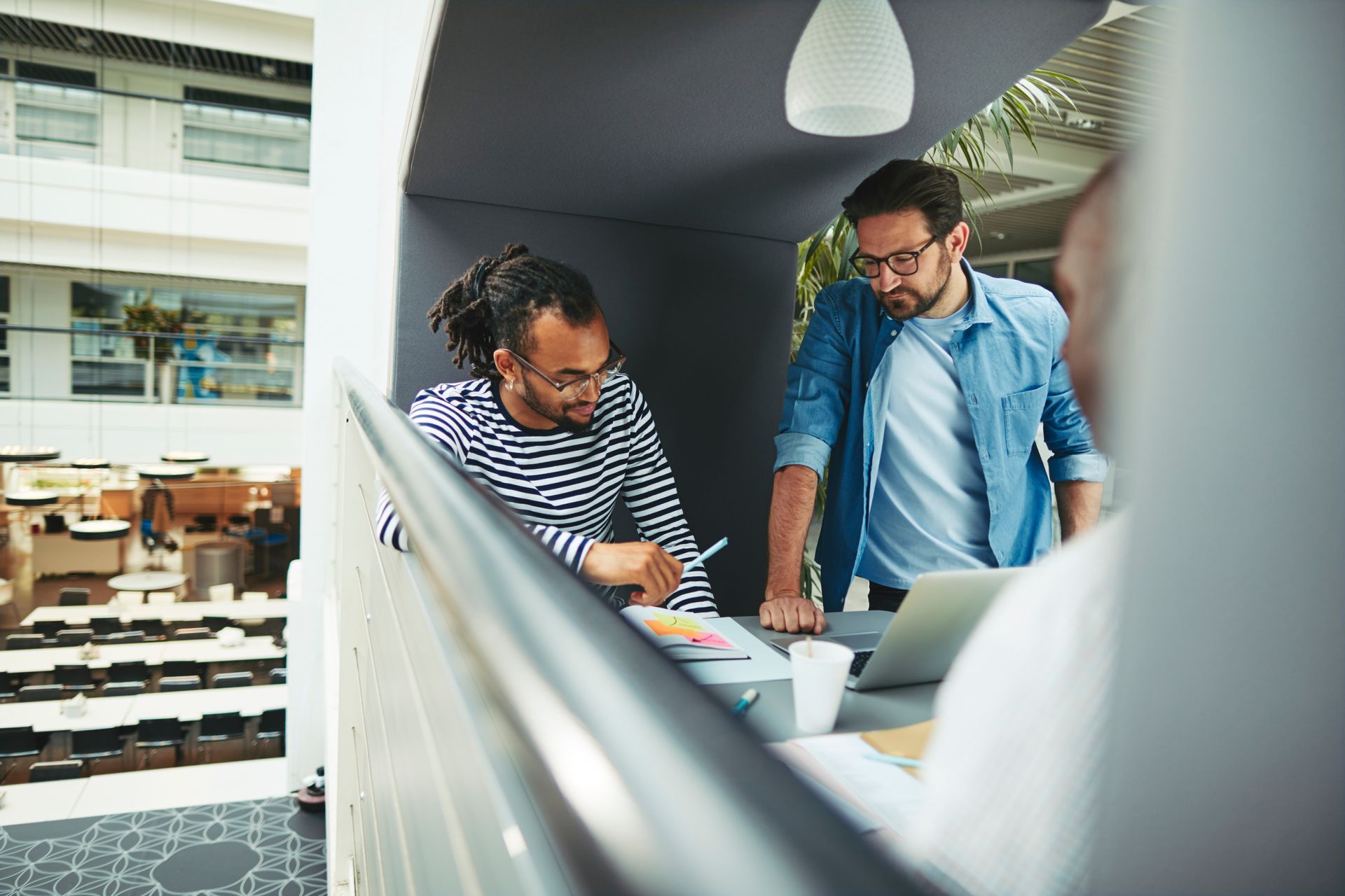 Two men collaborating, leaning on a railing with laptop, looking at documents. Modern office setting.