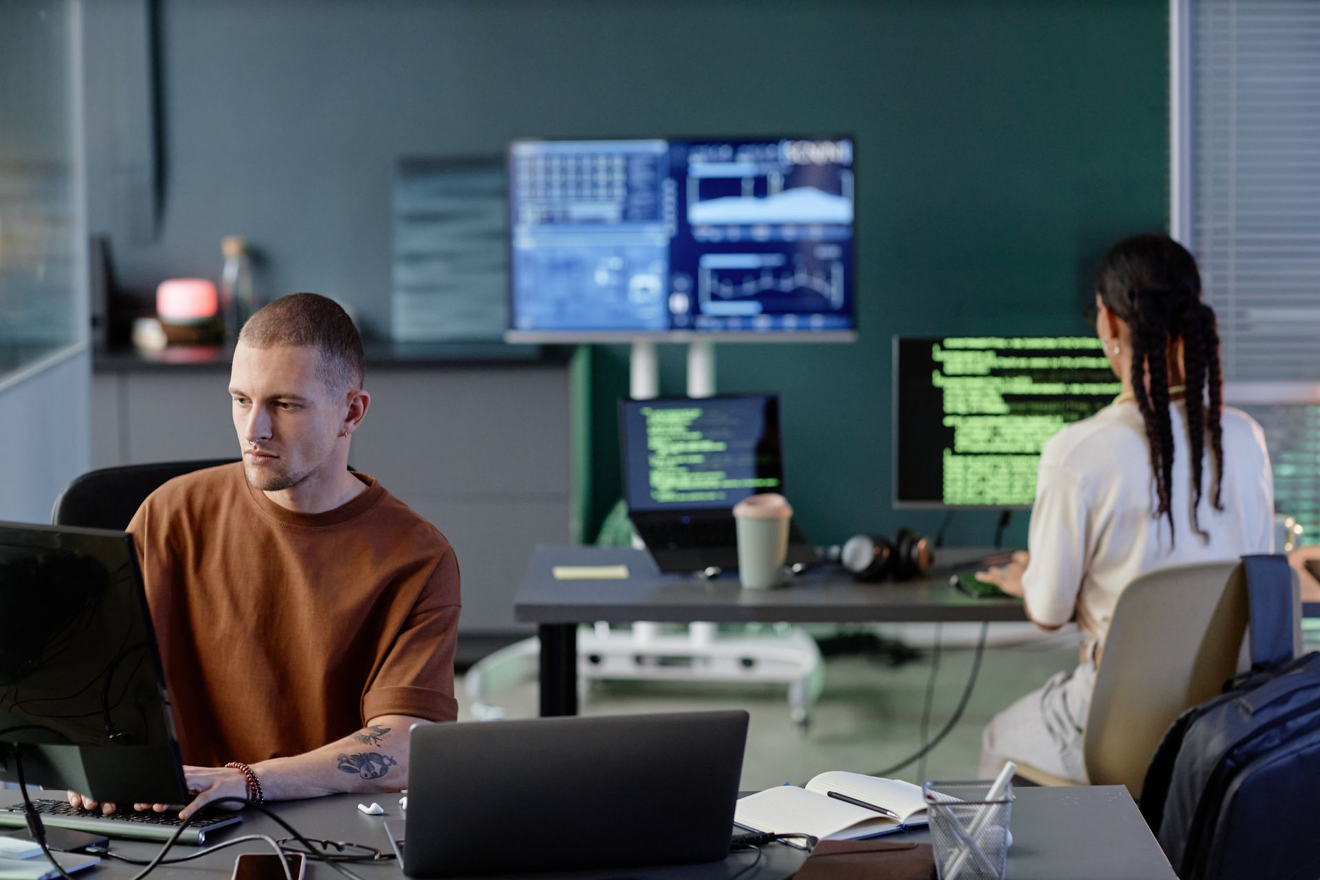 Two people working on computers in an office with multiple screens displaying code and data.
