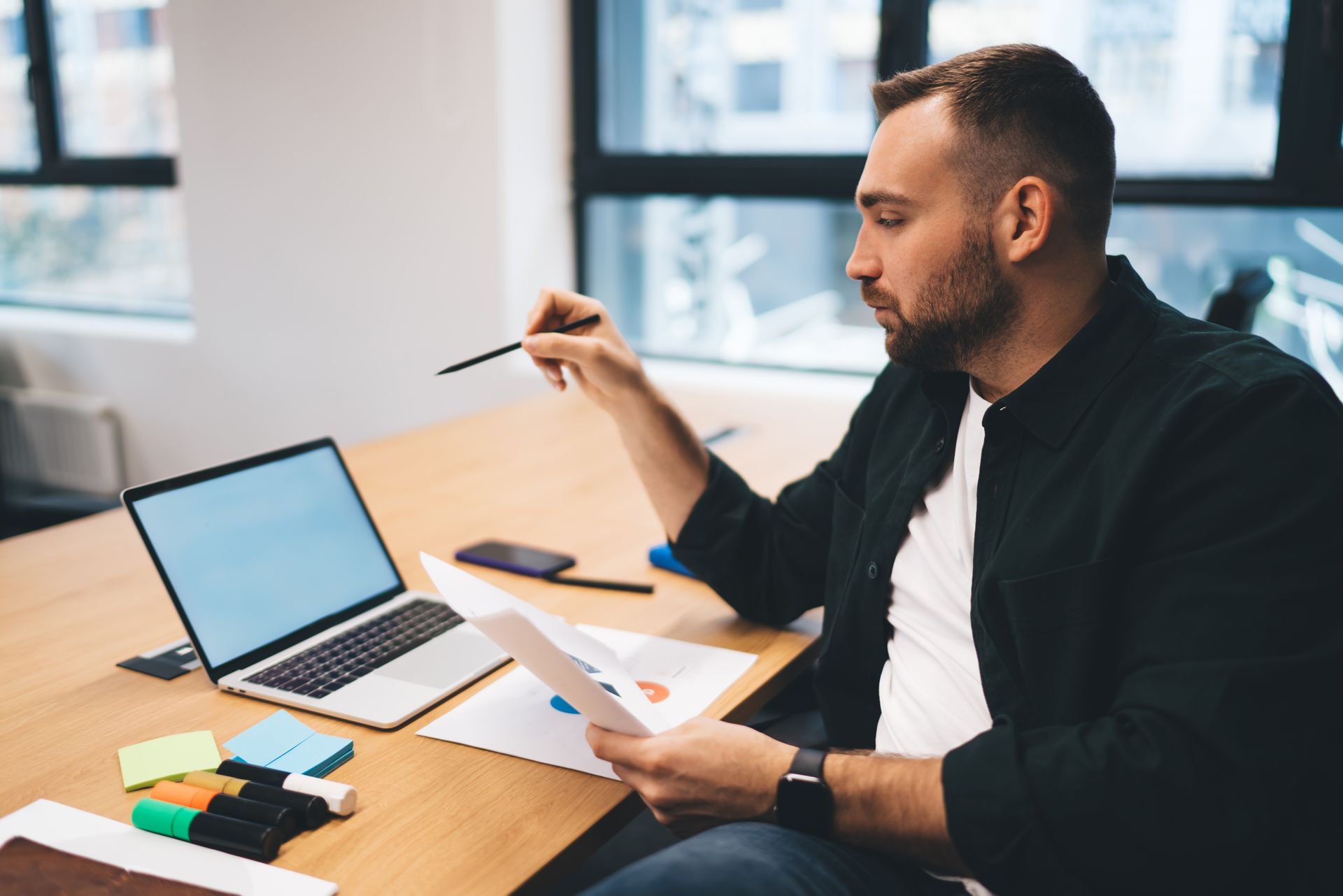 Man in a dark shirt looking at papers, pointing with a pen. Laptop and sticky notes on the table.