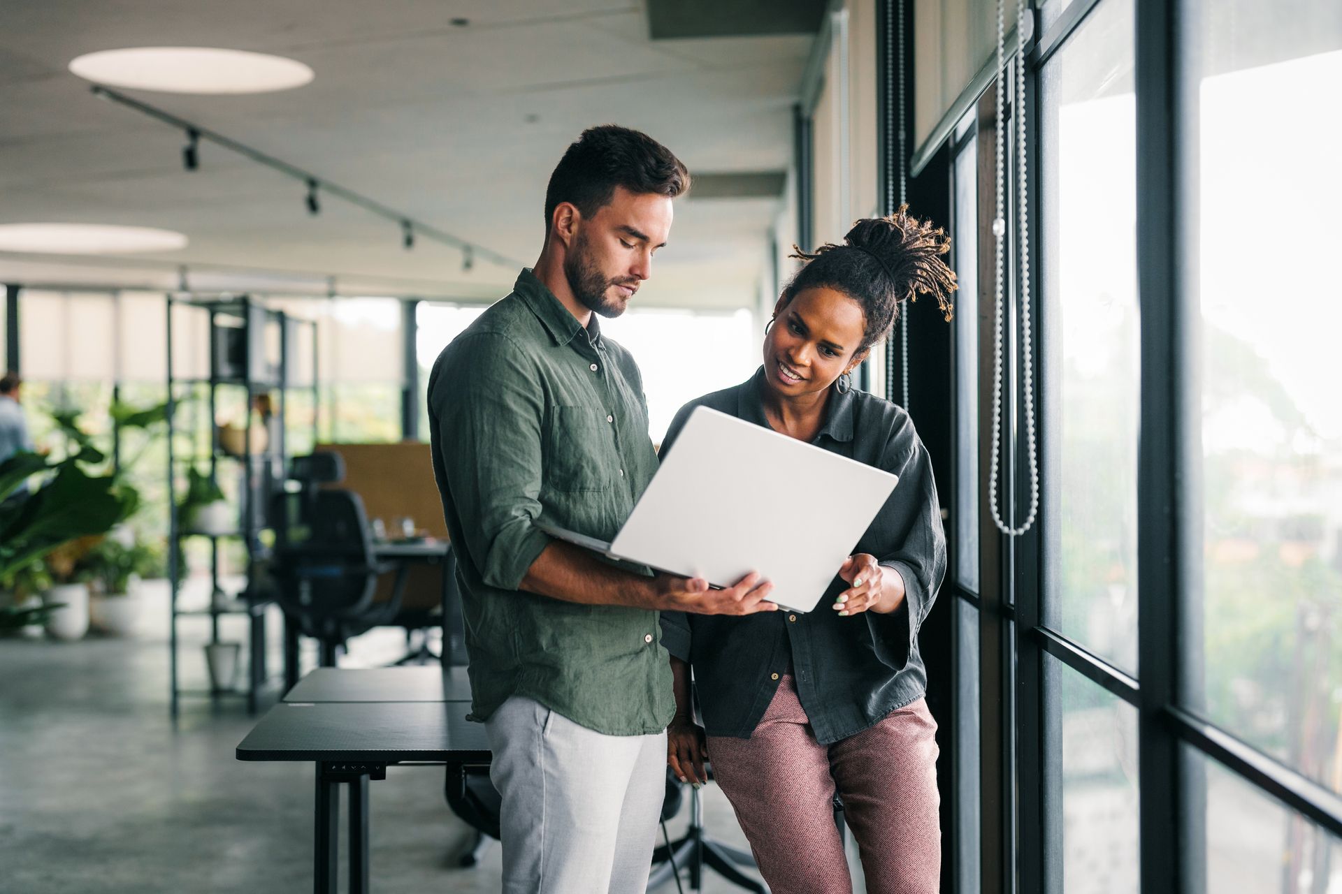 Two people looking at a laptop in an office setting. They stand by a large window.