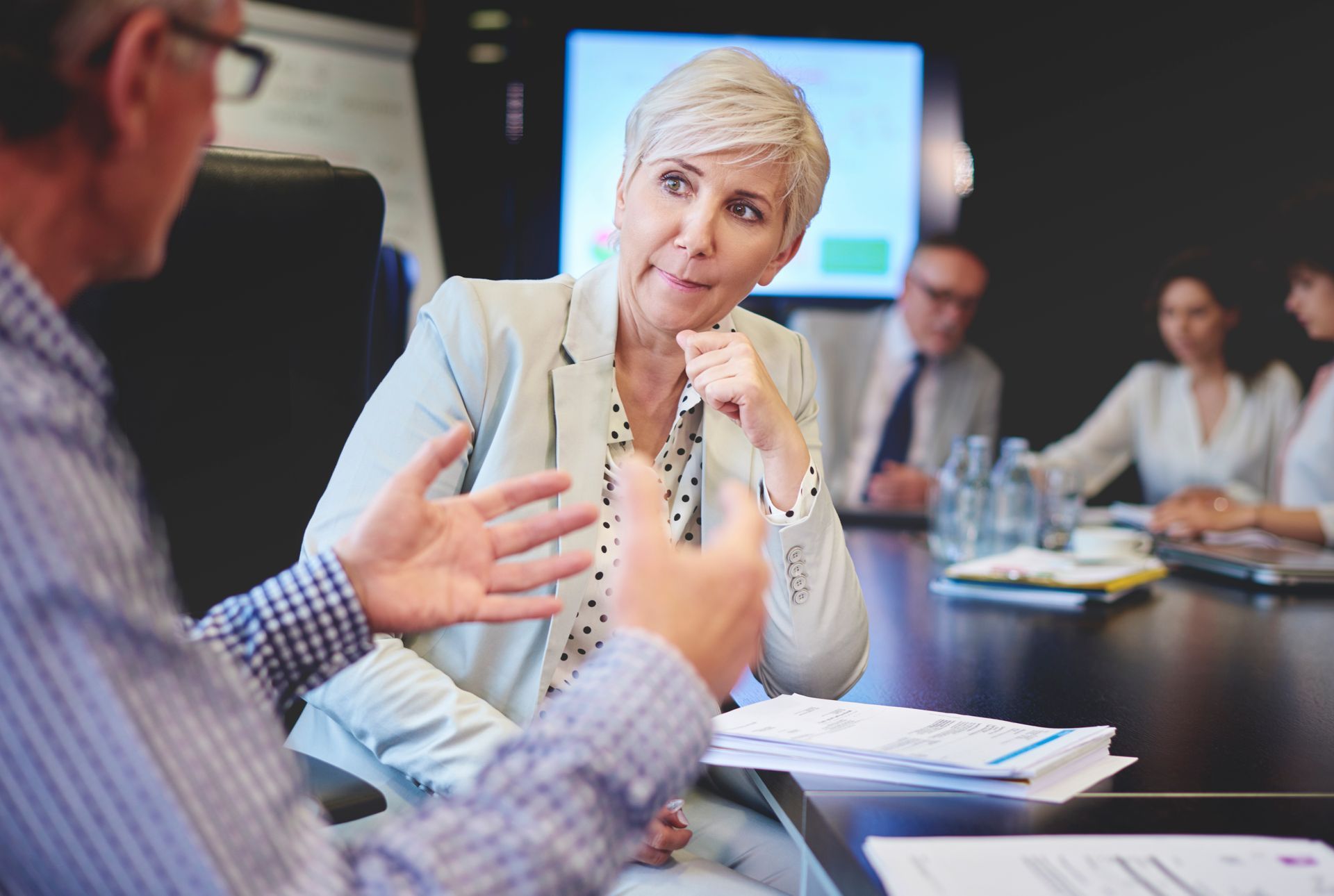 Professional colleagues in a boardroom setting, with one person gesturing while talking to a colleague across the table.