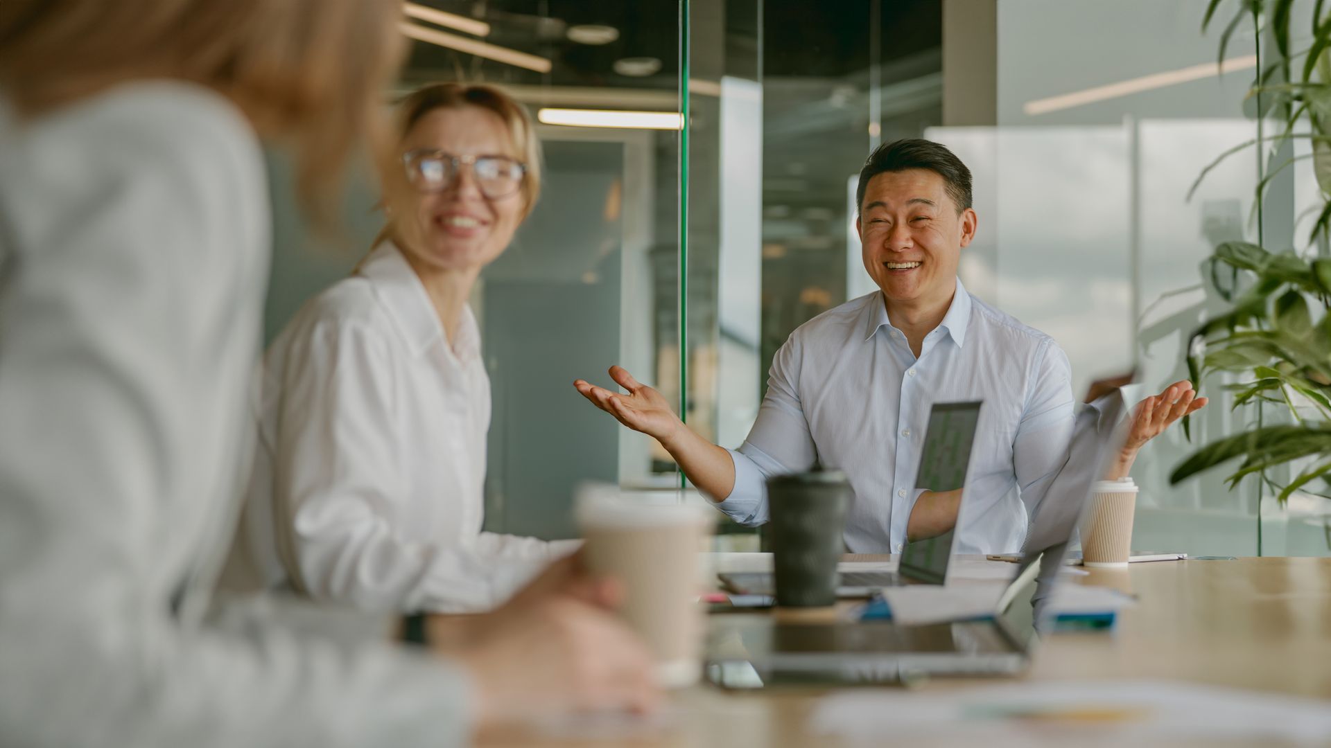 Three professionals in a bright office meeting, one speaking with open hands while others listen with smiling expressions.