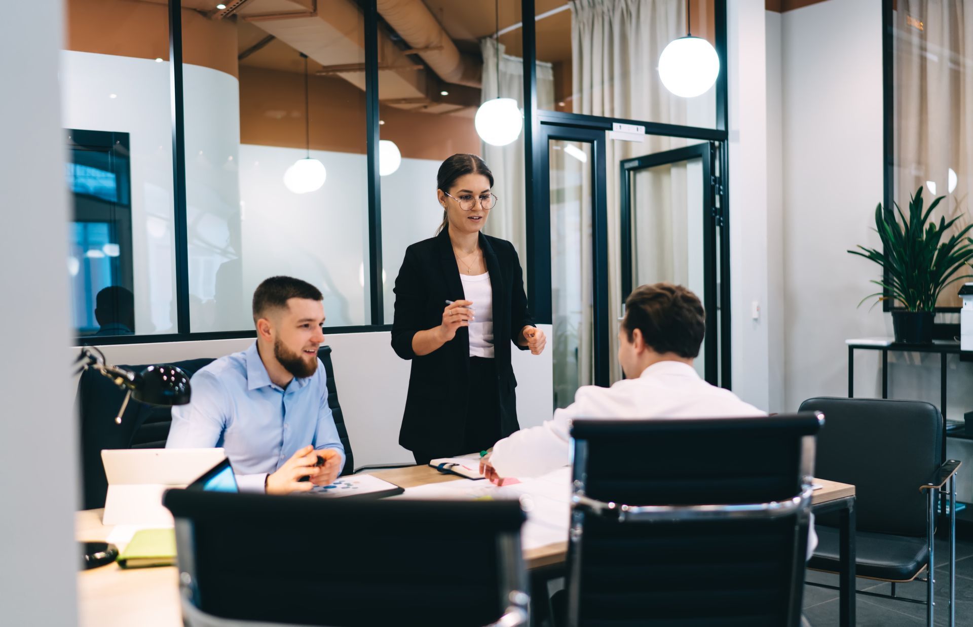 Businesswoman leading a meeting with two seated colleagues at a table in a modern office.