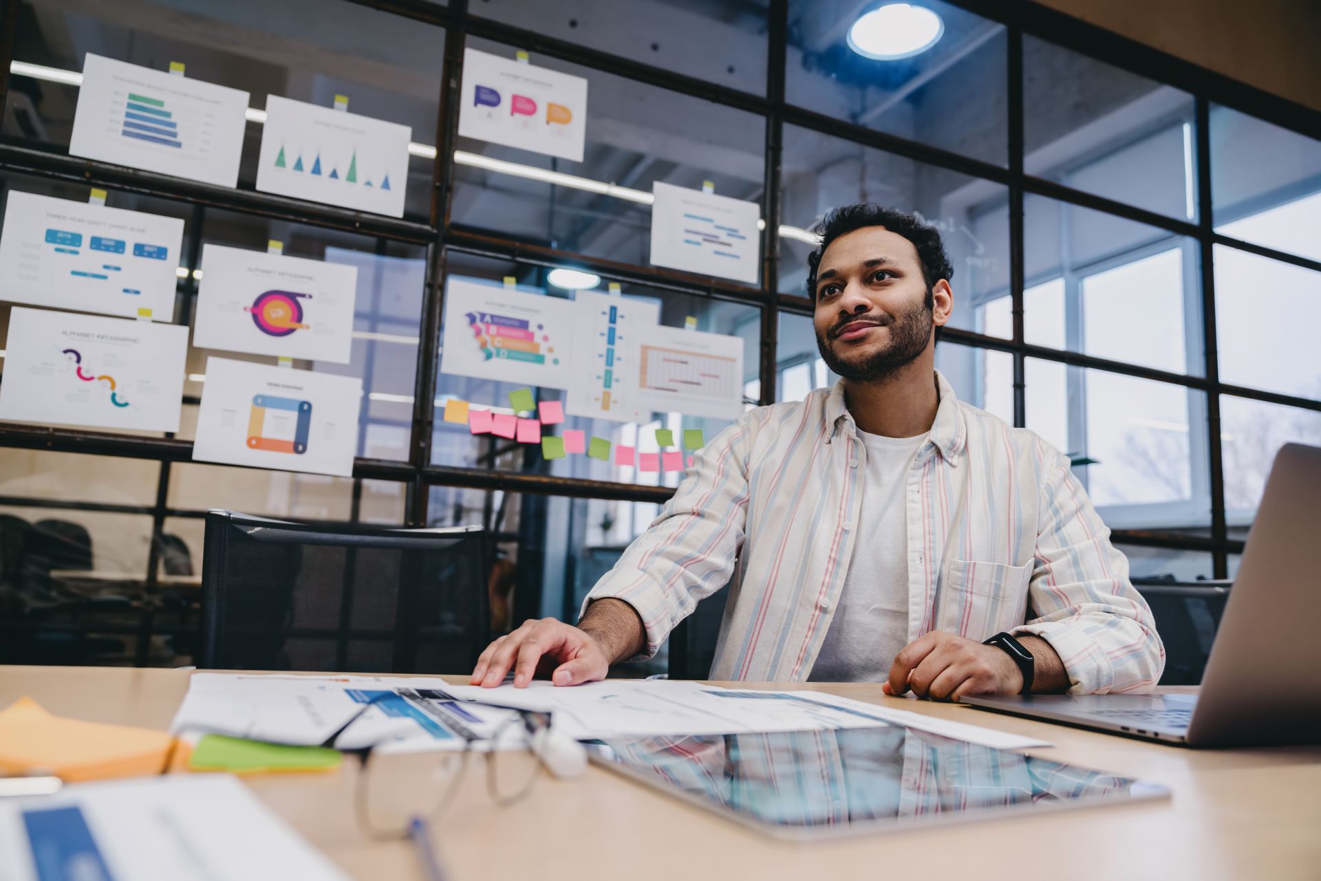 Man at desk smiles, looking at charts posted on glass wall. Laptop and documents present.