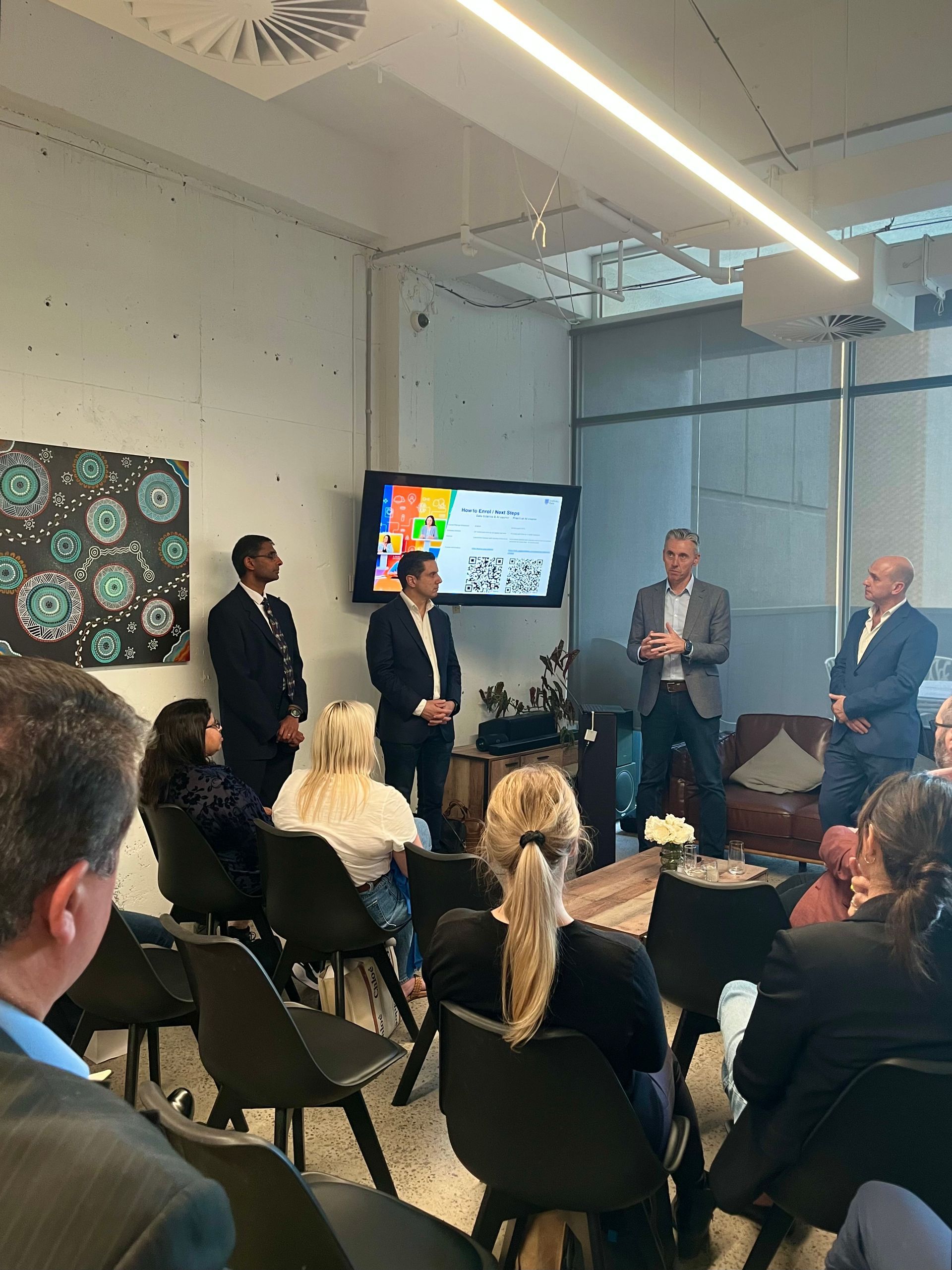 Four professionals in business attire speak to an audience in a modern office, standing before a screen with a presentation.