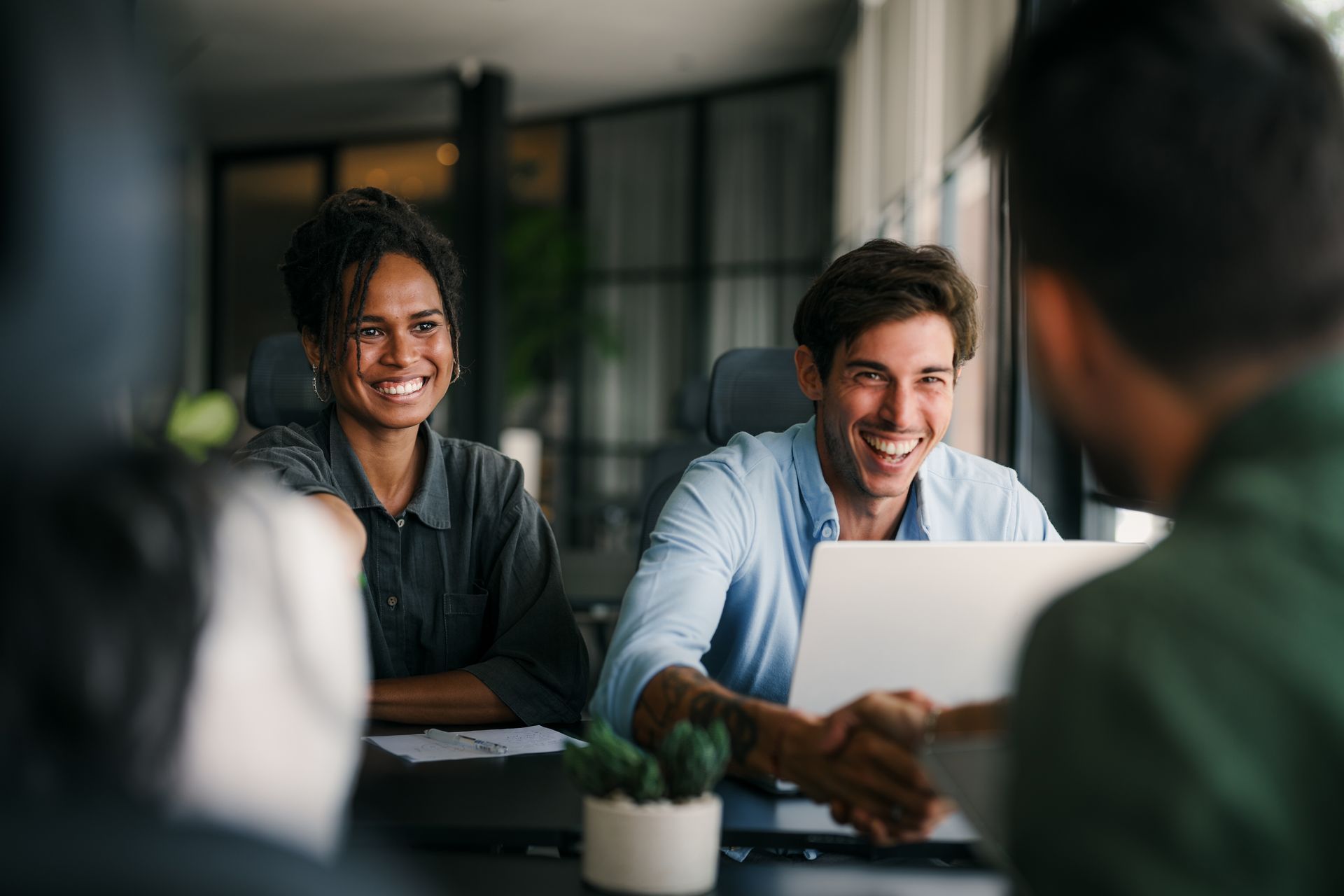 People in office shake hands, smiling. One holds laptop, plant on desk.