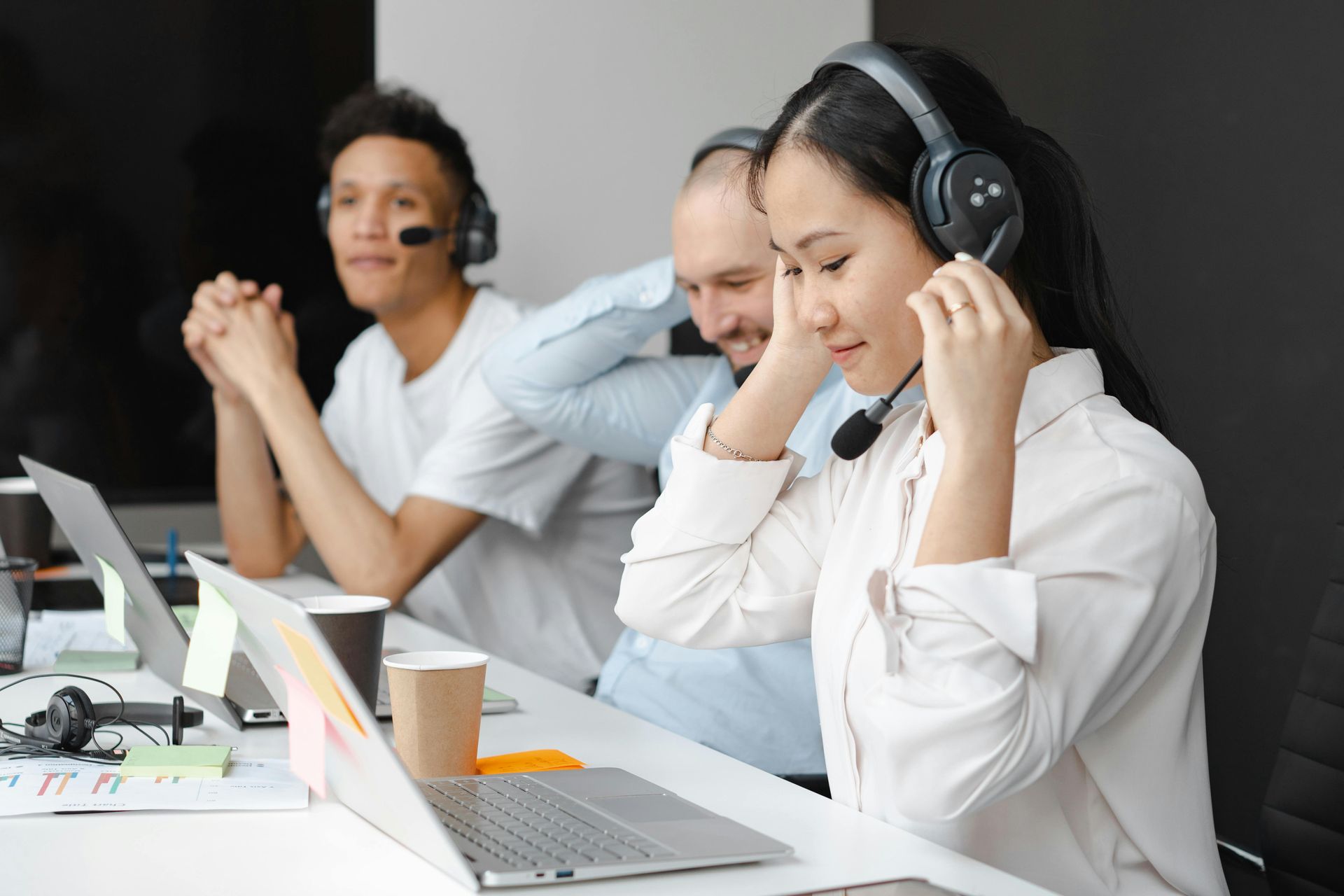Three people at desks wearing headsets, working on laptops in an office.