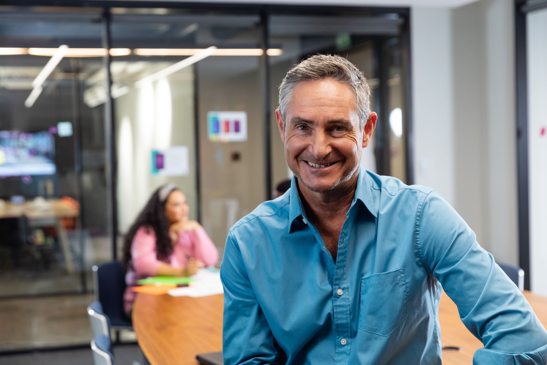A smiling person in a blue button-down shirt stands in a brightly lit office, with another person working in the background.