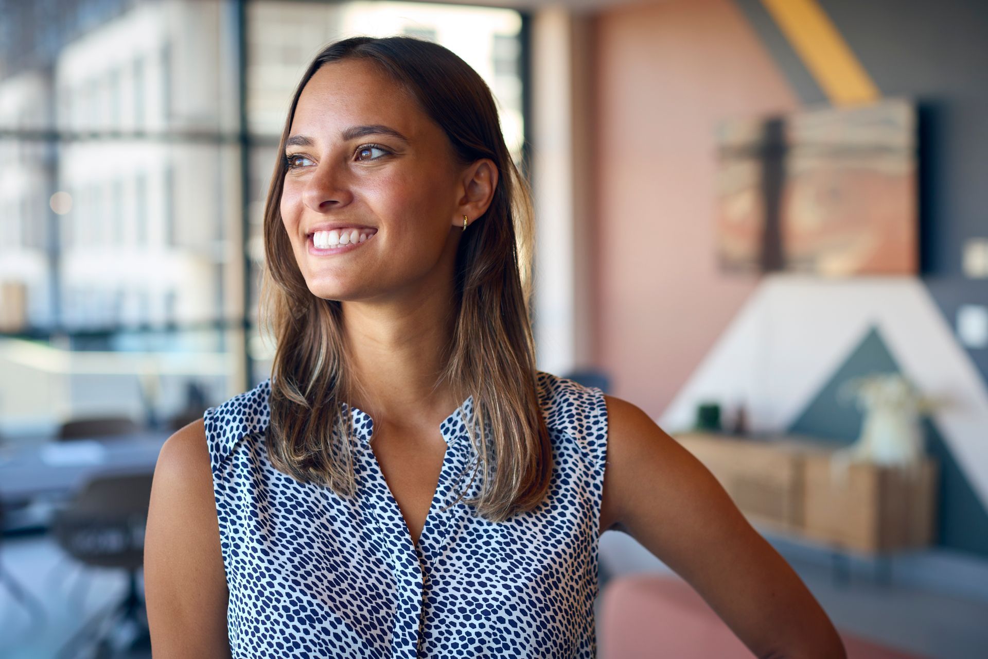 A person with long brown hair, wearing a patterned sleeveless top, smiles while looking away in a modern office.